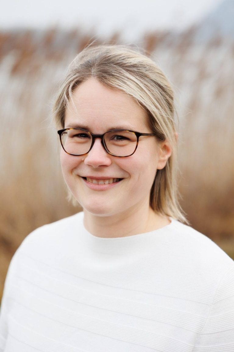 Junge Frau mit Brille, lächelnd im Freien vor einem unscharfen Hintergrund aus Pflanzen.