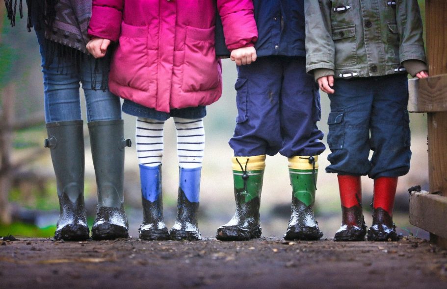 Vier Kinder in Gummistiefeln stehen Hand in Hand auf einem Holzsteg.