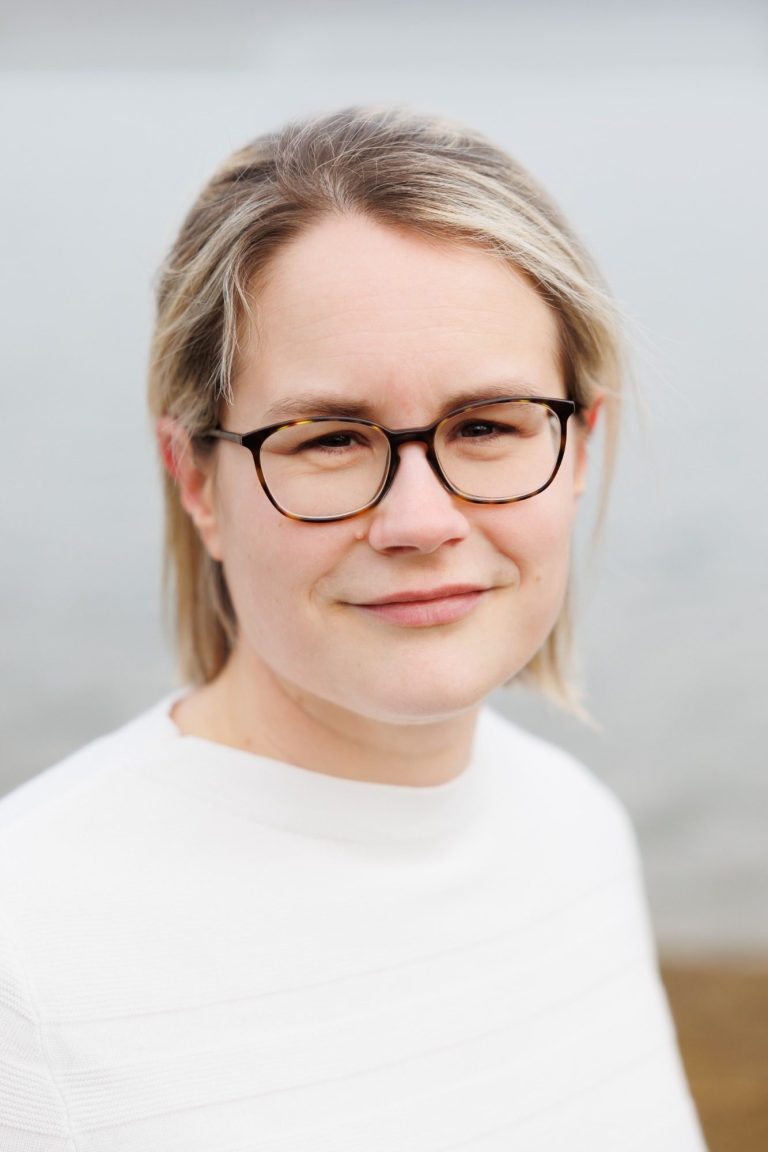 Portrait einer Frau mit Brille und blonden Haaren, lächelnd vor einem unscharfen Hintergrund.