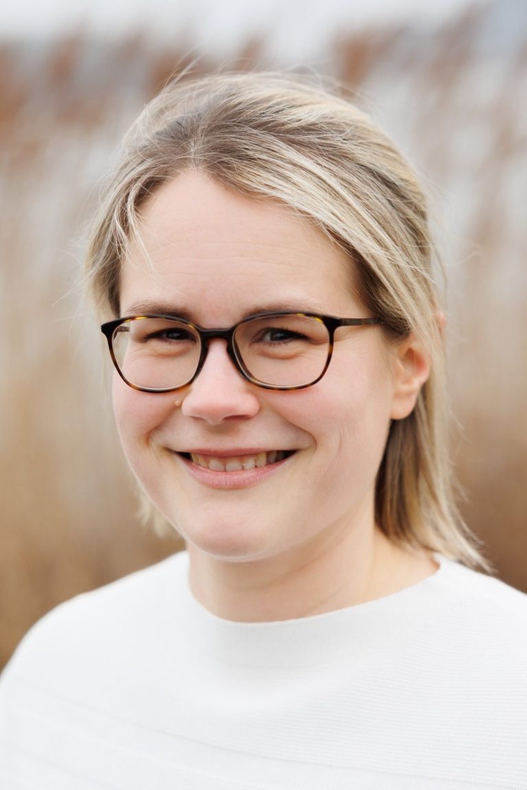 Foto von Huberta-Maria Schultes: Junge Frau mit Brille, lächelnd im Freien vor einem unscharfen Hintergrund aus Pflanzen.