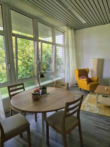 Heller Therapieraum mit einem runden Tisch, Stühlen und einem gelben Sessel. Pflanzen und Tageslicht.