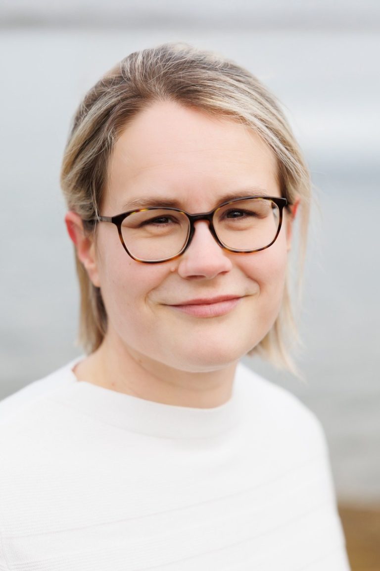 Portrait einer Frau mit Brille und blonden Haaren, lächelnd vor einem unscharfen Hintergrund.